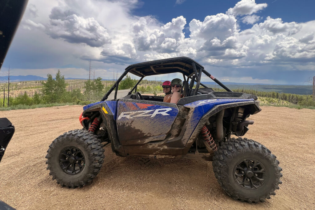 Two passengers wearing helmets inside a blue mud-splattered Polaris RZR XP 2-seater UTV rental on a dirt mountain trail in Colorado under a cloudy sky
