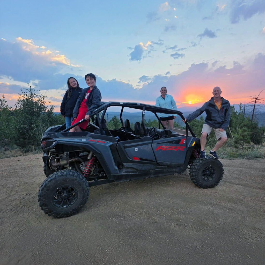 Family posing around a 4-seater Polaris RZR UTV rental at sunset on a scenic mountain trail near Woodland Park Colorado