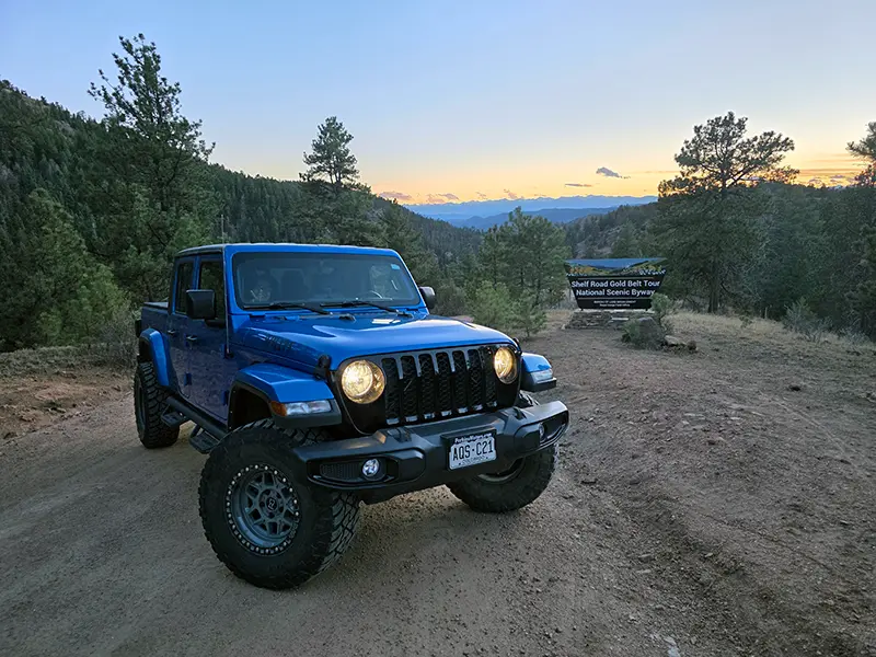 Colorado Jeep rental parked on a Woodland Park trail near the Shelf Road Gold Belt Tour sign during a Rocky Mountains adventure at sunset.