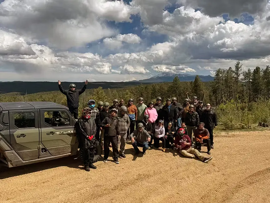 A large group of people wearing helmets posing outdoors on a dirt trail next to an off-road UTV, with pine forests and Pikes Peak visible in the background under a cloudy sky.