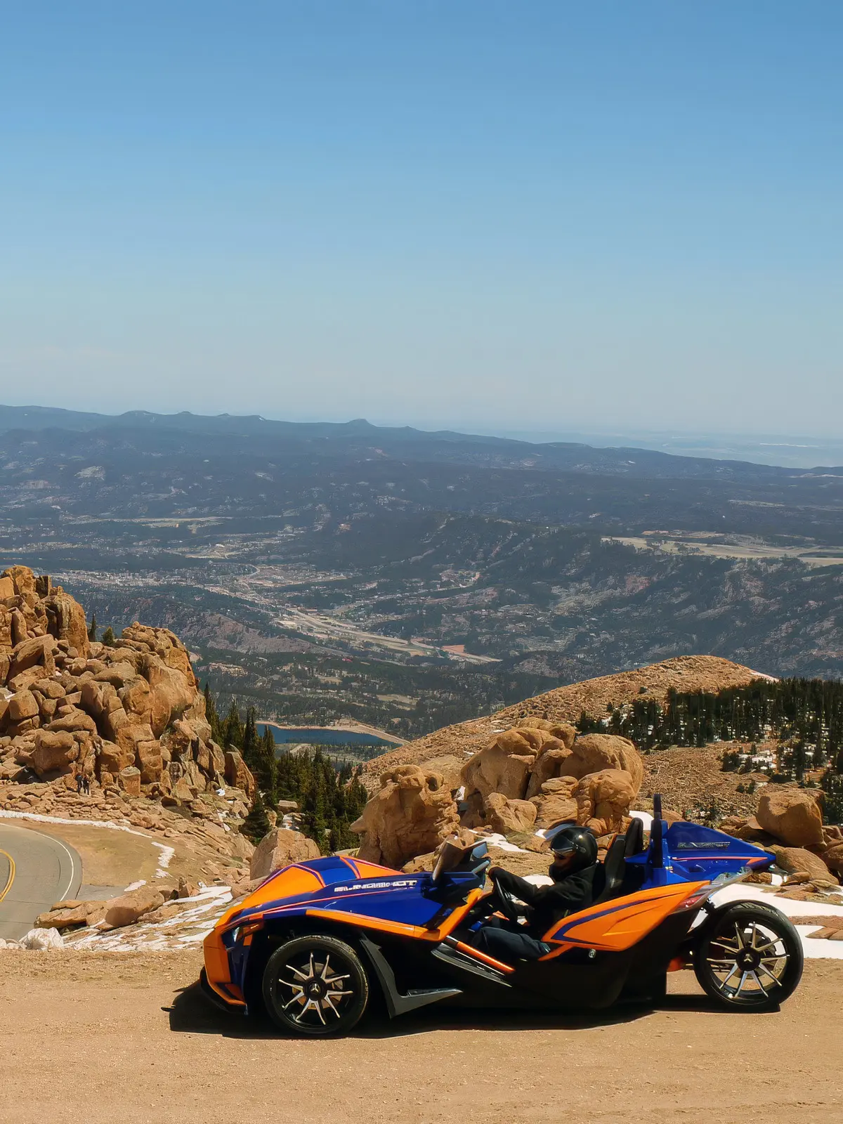 Blue and orange Polaris Slingshot rental parked on a scenic dirt overlook along Pikes Peak Highway with sweeping mountain views.